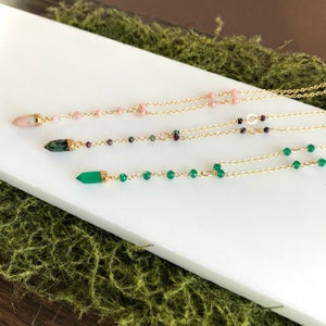 Op de afbeelding: Drie gouden kettingen met verschillende gekleurde edelsteen kralen en hangers. De bovenste ketting heeft roze kralen en een roze hanger. De middelste ketting heeft groene en paarse kralen en een groene hanger. De onderste ketting heeft groene kralen en een groene hanger.