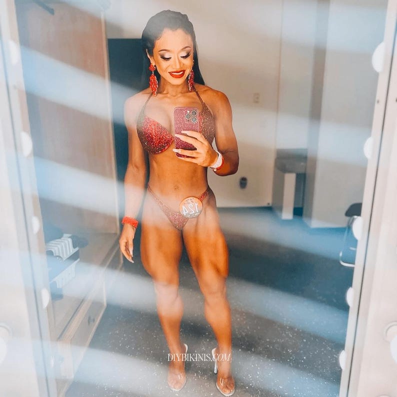 May include: A woman wearing a red sparkly bikini and a competition number. She is standing in front of a mirror and holding a phone.