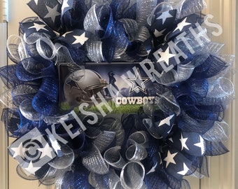 May include: A decorative wreath with a Dallas Cowboys theme. The wreath is made of blue, silver, and white mesh, with star-patterned ribbons. A central image displays the Cowboys logo and team name.