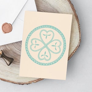 May include: A white envelope with a wax seal and a greeting card featuring a teal clover design, resting on a wooden slice. A small, antique-style stamp is also present. The card has a light beige background.