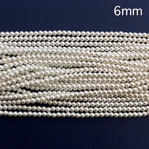 Könnte beinhalten: Nahaufnahme von Strängen mit 6 mm großen, weißen Kunstperlen vor dunkelblauem Hintergrund. Die Perlen sind gleichmäßig groß und in parallelen Linien angeordnet. Geeignet für die Schmuckherstellung.