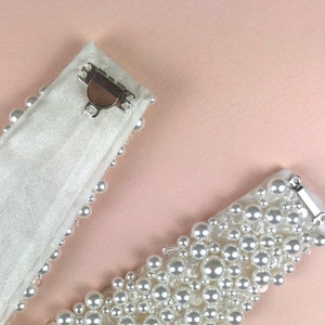May include: Two white satin ribbon belts with pearl embellishments and silver clasps. The belts are shown on a light pink background.