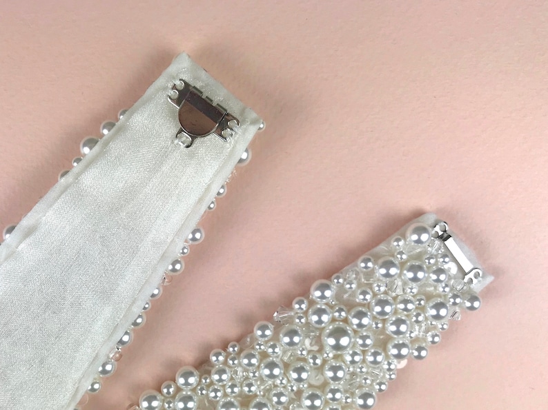 May include: Two white satin ribbon belts with pearl embellishments and silver clasps. The belts are shown on a light pink background.