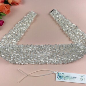 May include: A white pearl bridal sash with a satin ribbon tie. The sash is made of rows of pearls and is approximately 1.5 inches wide. The sash is perfect for adding a touch of elegance to a wedding dress.