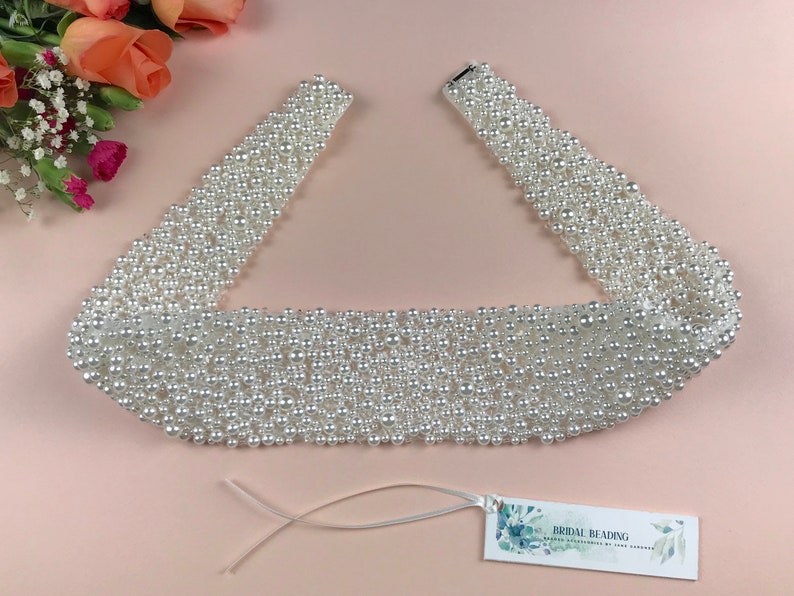 May include: A white pearl bridal sash with a satin ribbon tie. The sash is made of rows of pearls and is approximately 1.5 inches wide. The sash is perfect for adding a touch of elegance to a wedding dress.