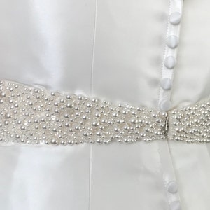 May include: A white satin dress with a pearl beaded belt. The belt is made of small, white pearls that are sewn together in a continuous line. The dress has a simple, elegant design.