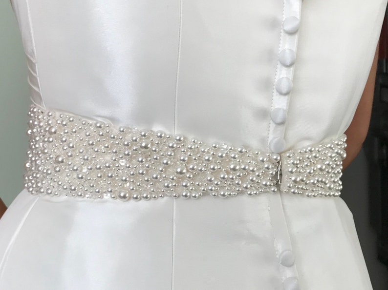 May include: A white satin dress with a pearl beaded belt. The belt is made of small, white pearls that are sewn together in a continuous line. The dress has a simple, elegant design.