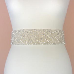 May include: A white beaded sash with a pearl-like finish. The sash is made of small, round beads that are closely packed together. It is a simple and elegant accessory that can be used to add a touch of sparkle to any outfit.