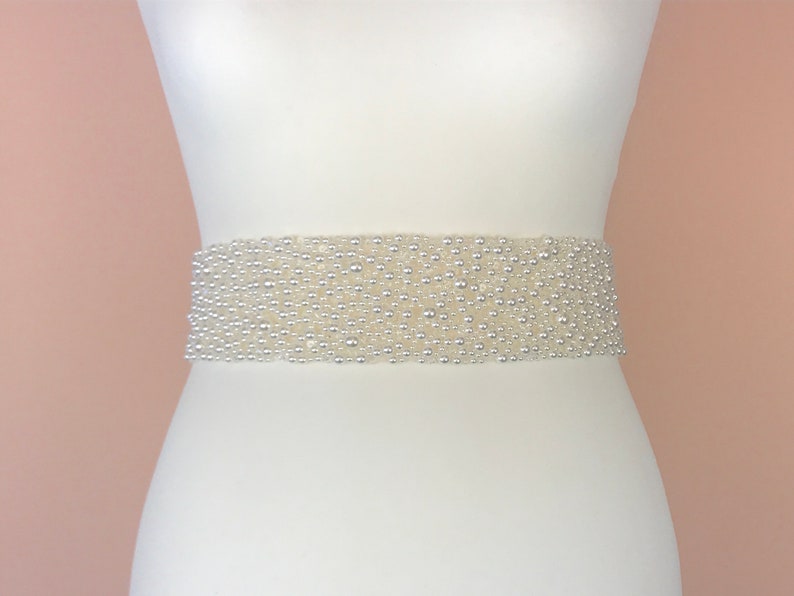 May include: A white beaded sash with a pearl-like finish. The sash is made of small, round beads that are closely packed together. It is a simple and elegant accessory that can be used to add a touch of sparkle to any outfit.