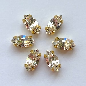 May include: Six clear marquise-shaped rhinestones with gold settings. The rhinestones are arranged in a circle on a white background.