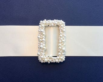 May include: A rectangular, pearl-covered frame sits on a white ribbon against a dark blue background. The frame is densely adorned with various sizes of white pearls, creating a textured, elegant border. The ribbon provides a contrasting, smooth, light backdrop.
