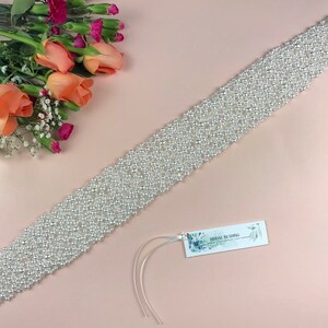May include: A white pearl bridal sash with a delicate, intricate design. The sash is made of small, round pearls that are sewn together in a pattern. The sash is perfect for adding a touch of elegance to a wedding dress.