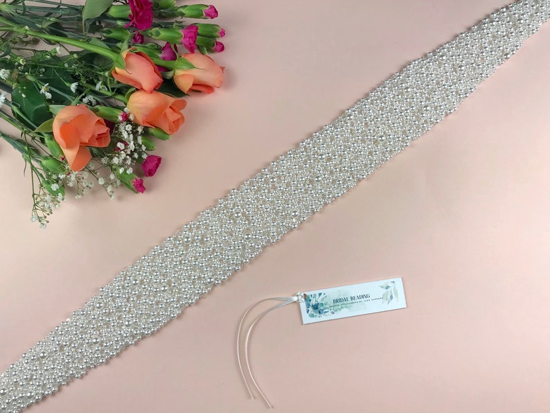 May include: A white pearl bridal sash with a delicate, intricate design. The sash is made of small, round pearls that are sewn together in a pattern. The sash is perfect for adding a touch of elegance to a wedding dress.