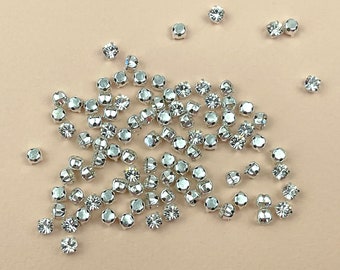 May include: A large group of silver-toned rhinestones with clear crystals. The rhinestones are scattered on a light brown surface.