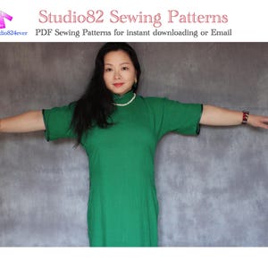 Può includere: Una donna indossa un vestito verde con un bordo nero e una collana di perle bianche. È in piedi davanti a uno sfondo grigio con le braccia distese.