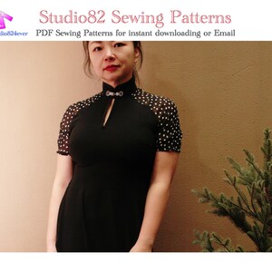 May include: A woman wearing a black dress with white polka dot sleeves and a mandarin collar. The dress has a keyhole opening at the neckline. The woman is standing in front of a brown wall with a small green plant to the right.