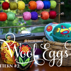 May include: Wool and Eggs pattern #2, a needlepoint embroidery pattern featuring a colorful floral design on a black egg-shaped pincushion. The pincushion is surrounded by other colorful egg-shaped pincushions, a wooden spool rack with colorful spools of thread, and a wicker basket.