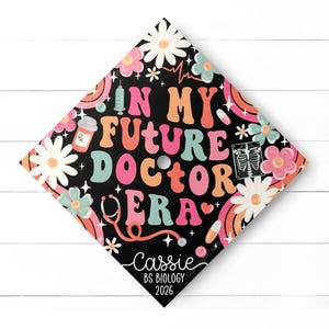 May include: A black graduation cap decorated with colorful text and graphics. The text reads "IN MY FUTURE DOCTOR ERA" in various colors. The cap also features flowers, rainbows, and medical-themed illustrations. The name "Cassie" and "BS Biology 2026" are also included.