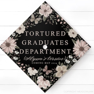 May include: Black graduation cap decorated with white and pink flowers, butterflies, and greenery. The text "Tortured Graduates Department (Alyssa's version) Coming May 2024" is printed on the cap.