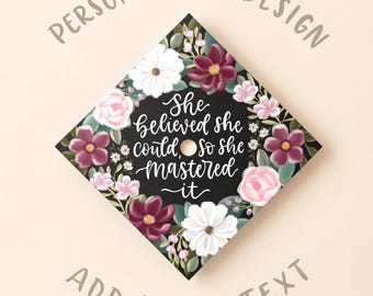 May include: A square graduation cap topper featuring a black background decorated with white, pink, and deep red floral illustrations. The center displays the white cursive text: She believed she could, so she mastered it. The words Personalized Design and Add Your Text appear above and below.