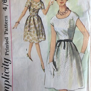 Könnte beinhalten: Vintage Simplicity Schnittmuster für ein Kleid, Größe 14, Brustumfang 86 cm. Das Schnittmuster zeigt zwei Illustrationen einer Frau, die ein Kleid mit rundem Ausschnitt und kurzen Ärmeln trägt. Das Kleid links ist mit einem Blumendruck, das Kleid rechts ist einfarbig weiß mit einer schwarzen Schärpe.