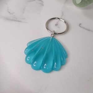 Op de afbeelding: Een turquoise blauwe schelp sleutelhanger met een zilveren ring. De schelp heeft een getextureerd oppervlak en een glanzende afwerking.