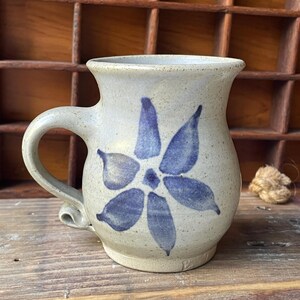 Peut inclure: Une tasse en céramique beige mouchetée avec un bord évasé et une anse incurvée. L'avant de la tasse présente un motif de fleur bleue à six pétales peint à la main. La base de la tasse possède un petit élément décoratif enroulé près de l'anse.