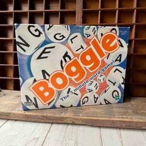 Op de afbeelding: Boggle woordspeldoos met witte dobbelstenen met letters. De doos heeft een blauwe achtergrond met het woord "Boggle" in oranje. De tekst "The 3 minute word game!" is ook zichtbaar.