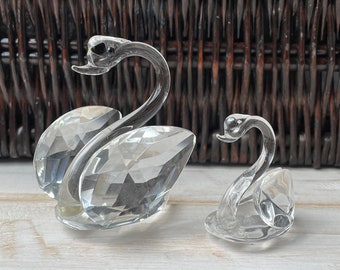 May include: Two crystal swan figurines, one larger than the other, with a clear, faceted design. The swans are positioned on a white surface.