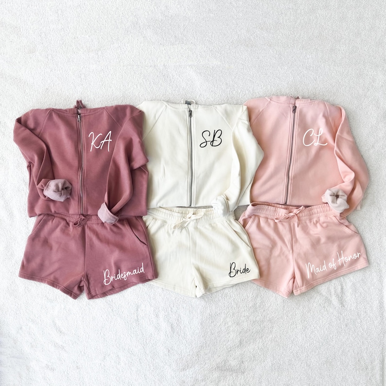 K&ouml;nnte beinhalten: Drei passende Sets aus Sweatshirts mit Rei&szlig;verschluss und Shorts in Mauve, Creme und Rosa. Das mauvefarbene Set hat "KA" auf dem Sweatshirt und "Bridesmaid" auf den Shorts. Das cremefarbene Set hat "SB" und "Bride". Das rosafarbene Set hat "CL" und "Maid of Honor".