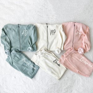 K&ouml;nnte beinhalten: Passende Sets aus Sweatshirts mit Rei&szlig;verschluss und Jogginghosen in Salbeigr&uuml;n, Creme und Ros&eacute;. Jedes Sweatshirt hat ein Monogramm, und die Jogginghosen sind mit "Maid of Honor", "Bride" und "Bridesmaid" beschriftet.