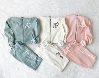 May include: Matching sets of zip-up sweatshirts and sweatpants in three colors: sage green, cream, and blush pink. Each sweatshirt has a monogram and each sweatpant has a title: "Maid of Honor", "Bride", and "Bridesmaid".