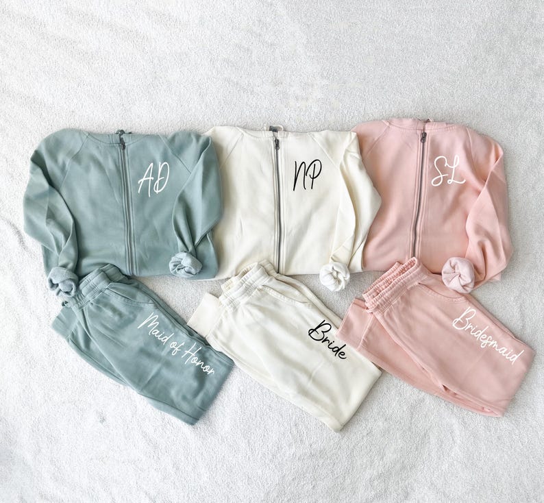 K&ouml;nnte beinhalten: Passende Sets aus Sweatshirts mit Rei&szlig;verschluss und Jogginghosen in Salbeigr&uuml;n, Creme und Ros&eacute;. Jedes Sweatshirt hat ein Monogramm, und die Jogginghosen sind mit "Maid of Honor", "Bride" und "Bridesmaid" beschriftet.