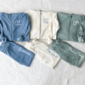 K&ouml;nnte beinhalten: Eine Kollektion passender Sweatshirts mit Rei&szlig;verschluss und Jogginghosen in Blau, Creme und Gr&uuml;n. Die Sweatshirts haben gestickte Initialen, und die Jogginghosen sind mit "Bridesmaid", "Mrs. Smith" und "Maid of Honor" beschriftet.
