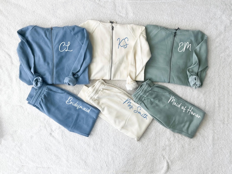 K&ouml;nnte beinhalten: Eine Kollektion passender Sweatshirts mit Rei&szlig;verschluss und Jogginghosen in Blau, Creme und Gr&uuml;n. Die Sweatshirts haben gestickte Initialen, und die Jogginghosen sind mit "Bridesmaid", "Mrs. Smith" und "Maid of Honor" beschriftet.