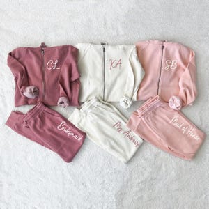 K&ouml;nnte beinhalten: Passende Sets aus Sweatshirts mit Rei&szlig;verschluss und Jogginghosen in Mauve, Wei&szlig; und Hellrosa. Die Sweatshirts haben gestickte Initialen, und die Jogginghosen sind mit "Bridesmaid", "Mrs. Andrews" und "Maid of Honor" beschriftet.
