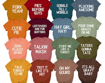 May include: A collection of colorful t-shirts with various Thanksgiving-themed phrases in white block letters. The shirts are in shades of orange, red, green, and brown, with phrases like "Fork Yeah!" and "Gobble Till I Wobble."