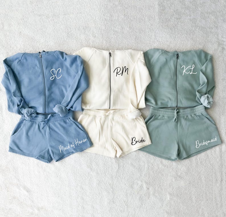 K&ouml;nnte beinhalten: Drei passende Loungewear-Sets in Blau, Creme und Gr&uuml;n. Jedes Set enth&auml;lt eine Jacke mit Rei&szlig;verschluss und Initialen sowie Shorts mit Text. Das blaue Set tr&auml;gt die Aufschrift "Maid of Honor", das cremefarbene Set "Bride" und das gr&uuml;ne Set "Bridesmaid".