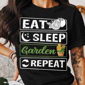 May include: Black t-shirt with white text that reads "Eat Sleep Garden Repeat". The text is in a blocky font. There is a crescent moon and stars above the word "Sleep". A tomato and a potted plant with yellow flowers are above the word "Garden".