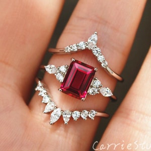 Peut inclure: Trois bagues en argent serties de pierres précieuses claires. La bague centrale présente une grande pierre rouge carrée. Les deux autres bagues sont incurvées et serties de petites pierres précieuses claires.