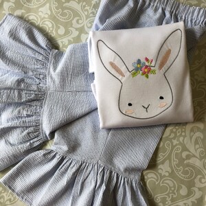 May include: A white and blue striped ruffle top with a white embroidered bunny with a floral crown. The bunny has pink ears and a pink nose.