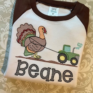 May include: A white and brown long-sleeved shirt with a brown turkey pulling a green tractor. The shirt has the name "Beane" embroidered on the front.