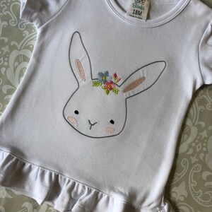 May include: White short-sleeved t-shirt with a ruffled hem. The shirt features an embroidered bunny with a floral crown. The tag reads "CRB Blanks 18M Made in China."