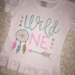 May include: White baby t-shirt with a pink, teal, and gold embroidered design that says "Wild One" with a dreamcatcher, feathers, and an arrow.