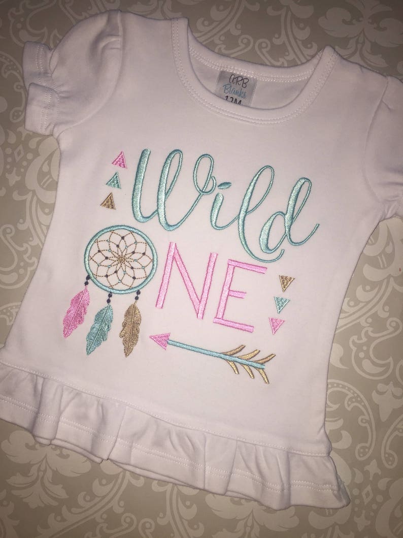 May include: White baby t-shirt with a pink, teal, and gold embroidered design that says "Wild One" with a dreamcatcher, feathers, and an arrow.