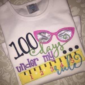 May include: White t-shirt with colorful embroidered text that reads "100 days under my rule" with a ruler and glasses graphic.