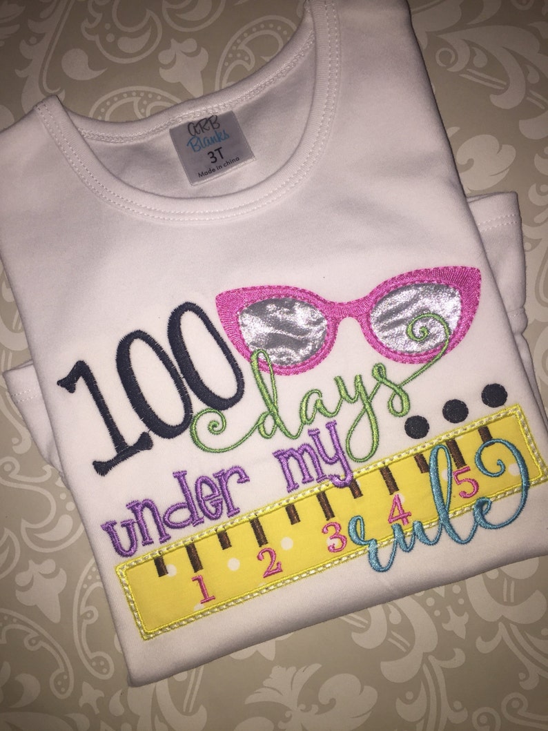May include: White t-shirt with colorful embroidered text that reads "100 days under my rule" with a ruler and glasses graphic.