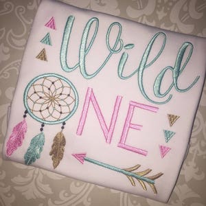 May include: White onesie with the text "Wild One" embroidered in turquoise and pink. The onesie also features a turquoise dreamcatcher, feathers, and an arrow.