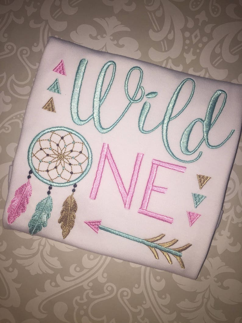 May include: White onesie with the text "Wild One" embroidered in turquoise and pink. The onesie also features a turquoise dreamcatcher, feathers, and an arrow.