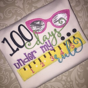 May include: A white shirt with embroidered text that reads "100 days under my rule" with a ruler and sunglasses graphic.